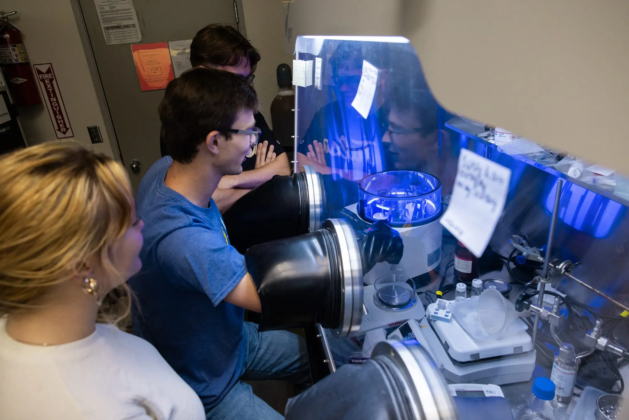 AMDG members working at an inert-atmosphere argon glovebox, one reaching into the gloves to handle a sample.